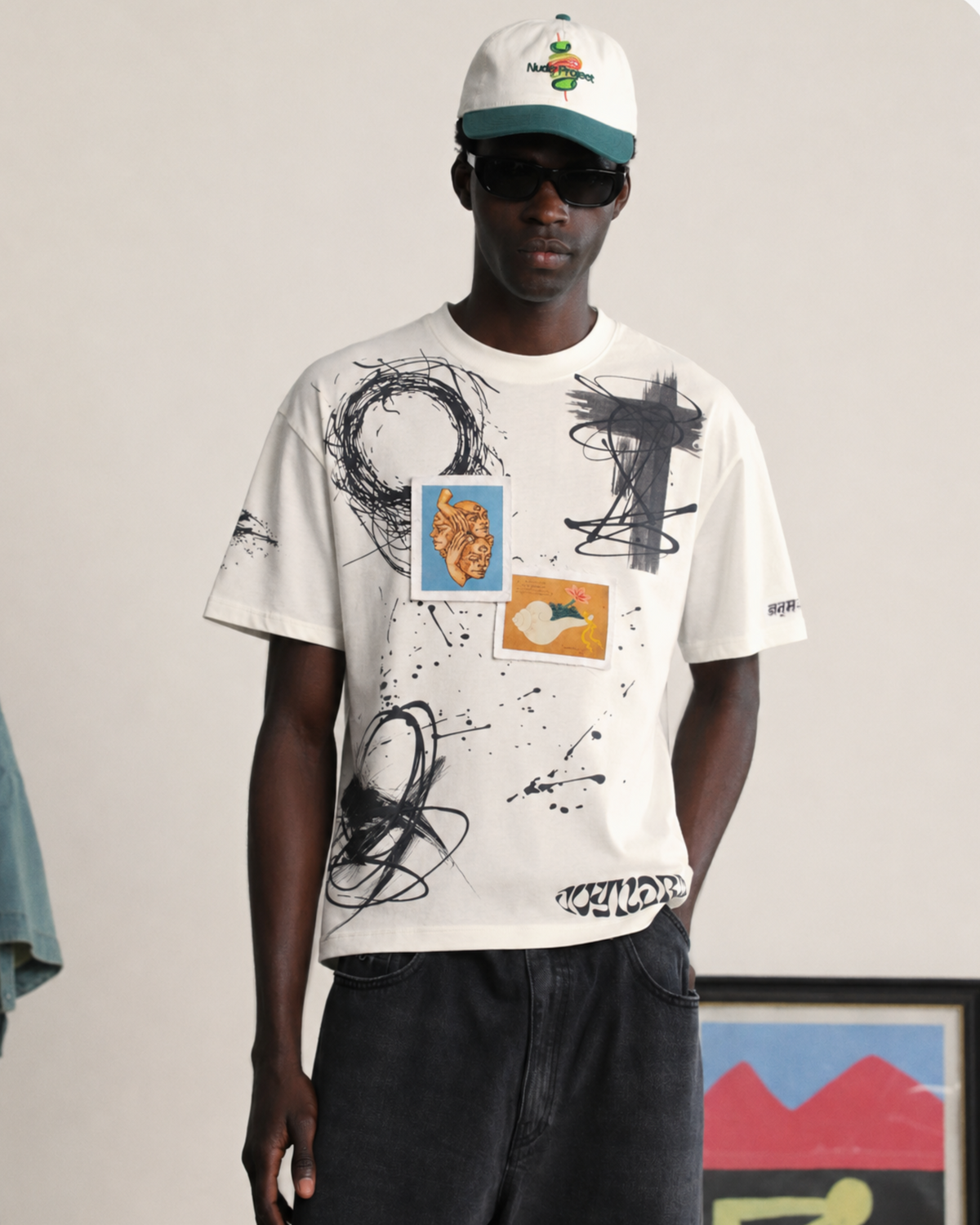 A model wearing a white graphic t-shirt with black scribble prints and colorful patchwork inserts, paired with dark jeans, sunglasses, and a white cap with a green brim.