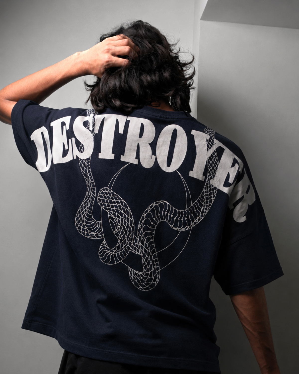 A person shown from behind wearing a dark navy blue oversized T-shirt with a large white graphic print reading 'DESTROYER' and a coiled snake design on the back, paired with black shorts against a plain white wall.