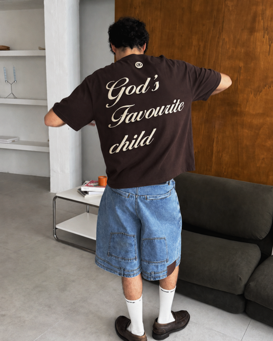 A man viewed from behind wearing an oversized dark brown t-shirt with 'God's Favourite child' printed in white script across the back, paired with denim shorts, white socks, and brown loafers in a modern room.