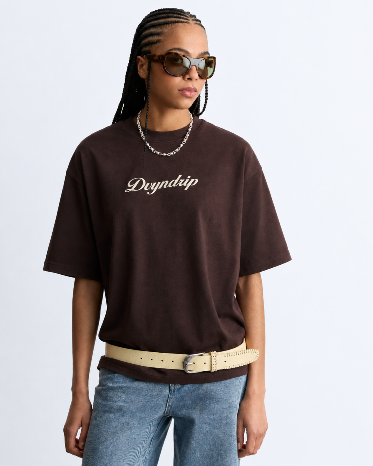 A woman modeling an oversized dark brown t-shirt with 'Dvyndrip' printed in white script across the chest, styled with light blue jeans, a cream belt, and sunglasses against a light gray background.