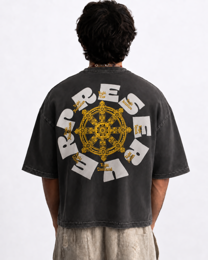 A person viewed from behind wearing a faded black graphic T-shirt with a large white and yellow 'PRESERVE' design and a central yellow wheel motif, paired with light beige pants against a plain white studio background.