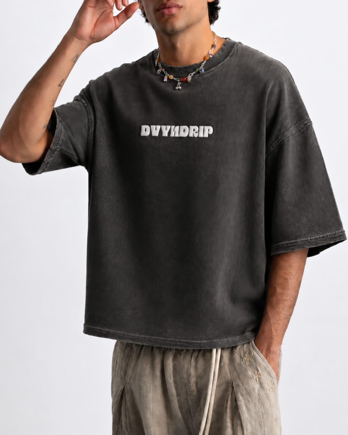 A man wearing a washed black graphic t-shirt with 'DVYNDRIP' printed in white across the chest, paired with beige drawstring pants, against a plain light gray background.
